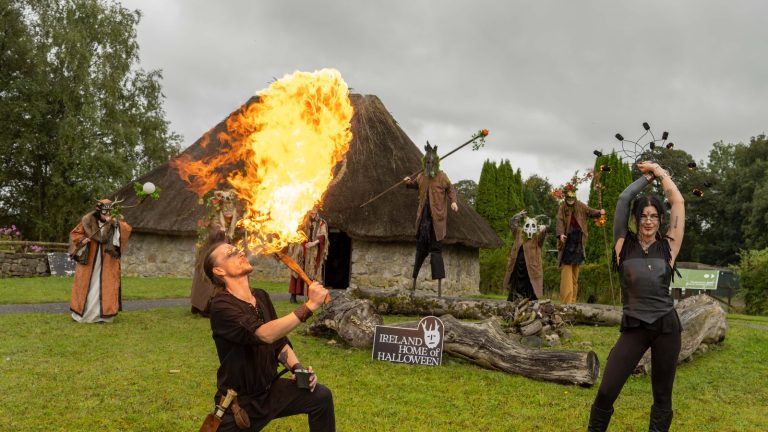 A fire performer breathes a huge flame in front of a thatched cottage at Dún na Sí, Moate, as costumed Samhain performers and a fire dancer look on.